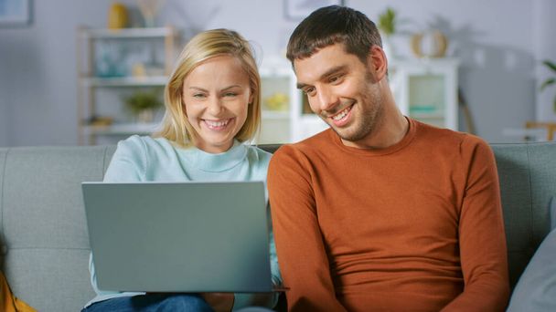 Cute Young Couple Sits on a Sofa at Home, They use Laptop Computer and Have Fun. Portrait of Beautiful Happy Young Couple in Love. - 写真・画像