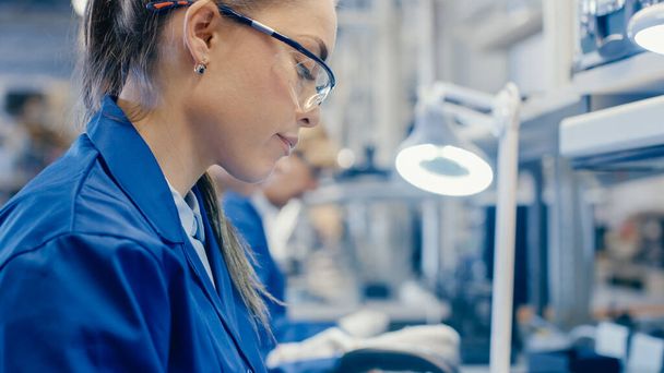 Woman Electronics Factory Worker in Blue Work Coat and Protective Glasses is Assembling Smartphones with Tweezers and Screwdriver. High Tech Factory Facility with more Employees in the Background. - Foto, Bild