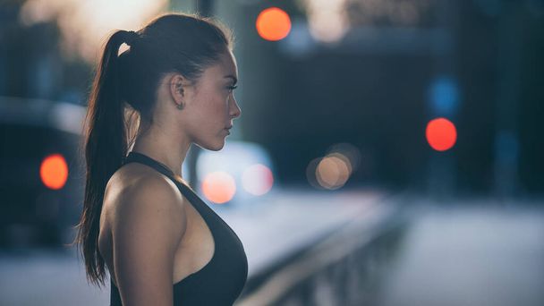 Portrait Shot of a Beautiful Confident Fitness Girl in Black Athletic Top on a Street. She is a Brunette with Brown Eyes with Her Hair Tied in an Ponytail. Shot is Taken in an Urban Environment. - Fotó, kép