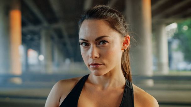 Portrait Shot of a Beautiful Confident Fitness Girl in Black Athletic Top on a Street. She is a Brunette with Brown Eyes with Her Hair Tied in an Ponytail. Shot is Taken in an Urban Environment. - Foto, Bild