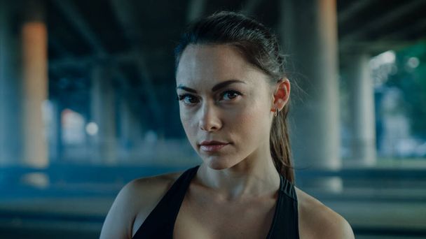 Portrait Shot of a Beautiful Confident Fitness Girl in Black Athletic Top on a Street. She is a Brunette with Brown Eyes with Her Hair Tied in an Ponytail. Shot is Taken in Dark Urban Environment. - Foto, afbeelding