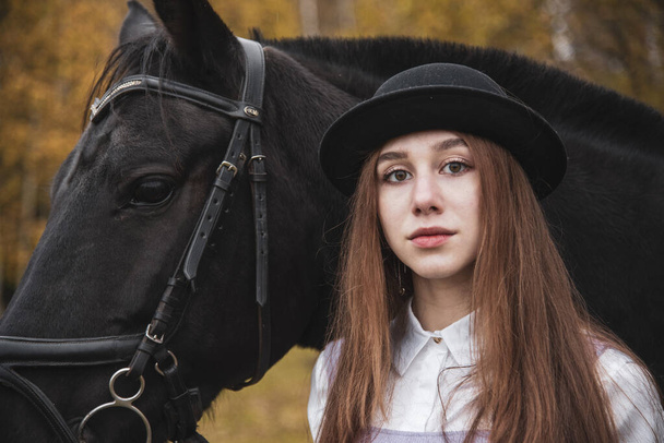 Red-haired girl in a black hat with a Chestnut horse in the autumn forest. - Foto, imagen