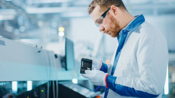 High-Tech Factory: Quality Control Engineer Picks up and Visually Checks Smart Electronic Device for Possible Damages. In the Background Assembly Line for PCB with Surface Mount Pick and Place - 写真・画像