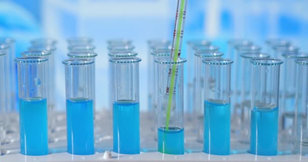 Ученый вводит химикаты в пробирки. Тестовая трубка цветной жидкости на лабораторном столе на заднем плане стеклянных колб. заливают из стеклянной пипетки в пробирку, освещенную голубым светом - Кадры, видео