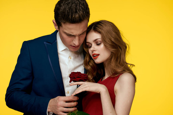 Un hombre romántico abraza a una mujer en un vestido rojo con una rosa en la mano sobre un fondo amarillo - Foto, imagen