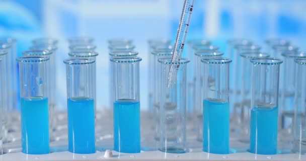 Wetenschapper injecteert chemicaliën in reageerbuizen. Testbuis met gekleurde vloeistof op de laboratoriumtafel op de achtergrond van de glazen kolven. gegoten van glazen pipet in reageerbuis verlicht met blauw licht - Video
