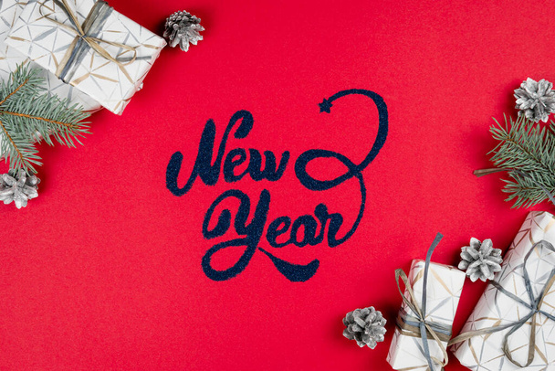 Újévi felirat piros háttér, fenyőfa ágak, ezüst kúp és ajándék doboz körül, lapos fektetés. Fogalma üdvözlő kártya. Felülnézet. - Fotó, kép