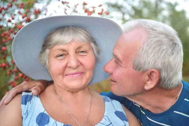 Αυθεντικό υπαίθριο πλάνο του ηλικιωμένου ζευγαριού που διασκεδάζει στον κήπο και είναι ευλογημένο με αγάπη. Κατά τη διάρκεια του παιχνιδιού τους άνθρωπος προσπαθεί να φιλήσει τη γυναίκα του και αυτή χαμογελά. Αγάπη και οικογενειακή έννοια. - Φωτογραφία, εικόνα