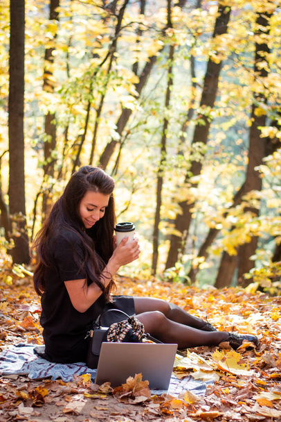 A brunette woman is sitting in an autumn park with a laptop and a cup of coffee. Student. Teacher. Online education. - Fotó, kép