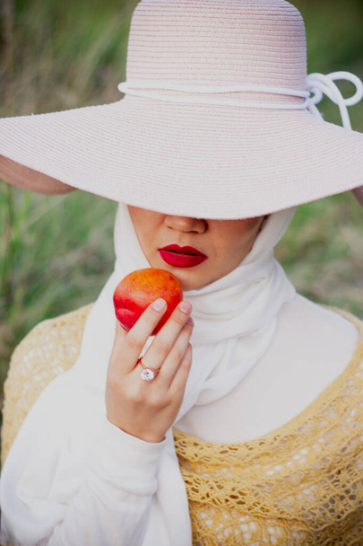 A Muslim woman in a hat sits on the field and eats peaches. Red lips. beauty - Foto, immagini