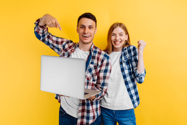 excited young couple, a man and a woman in plaid shirts, use a laptop and show a winning gesture, on a yellow background - Foto, Imagen