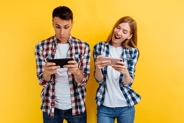 happy couple, a man and a woman in white t-shirts and plaid shirts, play video games on a smartphone, isolated yellow background - Foto, Imagem