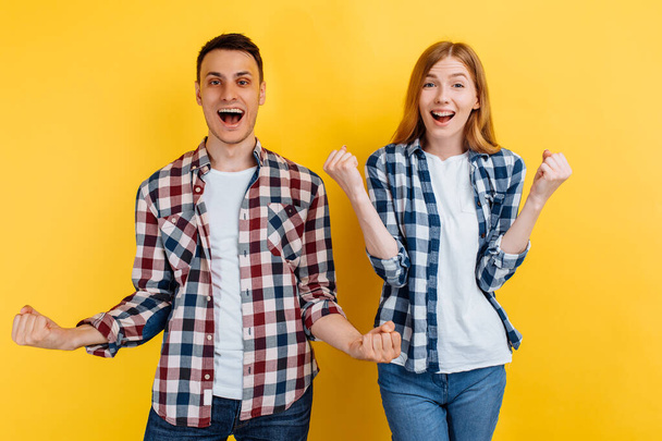 Shocked young man and woman, in checkered shirts and white t-shirts, celebrate the victory, showing a victory gesture, on a yellow background - Photo, image