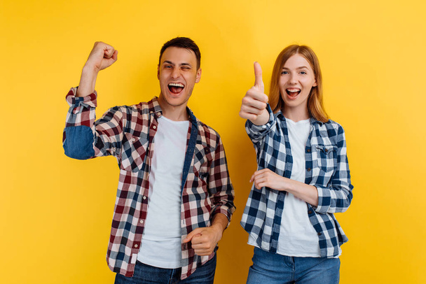 Shocked young man and woman, in checkered shirts and white t-shirts, celebrate the victory, showing a victory gesture, on a yellow background - Foto, afbeelding