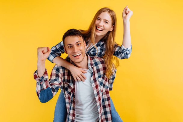 cheerful young couple, man and woman showing victory gesture, ride on back, on yellow background - Foto, immagini