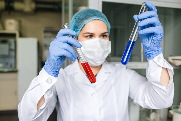 Young female scientist in medical cap and protective mask are doing investigations with test tubes red and blue solution in laboratory. - Photo, image