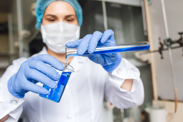 Jeune chercheuse en casquette médicale et masque de protection font des recherches avec une solution bleue de tube à essai en laboratoire. - Photo, image