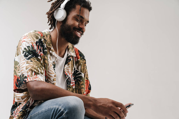 Image of a positive cheery african guy isolated over grey wall background using mobile phone while listening music with headphones - Foto, afbeelding