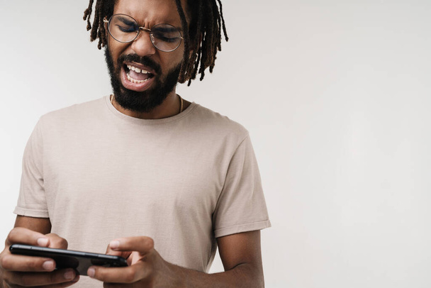 Photo of nervous emotional african guy playing games by mobile phone isolated over grey wall background in eyeglasses - Fotografie, Obrázek