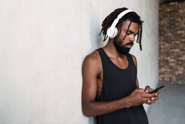 Imagen de un serio deportista afroamericano usando teléfonos celulares y auriculares en interiores - Foto, imagen