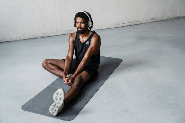 Image of strong african american sportsman in headphones doing exercise while working out on mat indoors - Fotó, kép