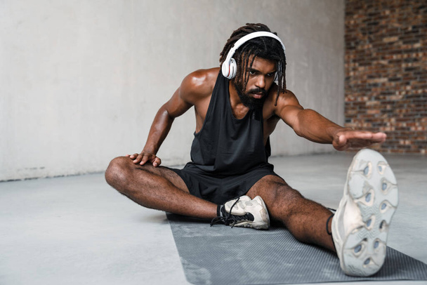 Image of strong african american sportsman in headphones doing exercise while working out on mat indoors - Zdjęcie, obraz