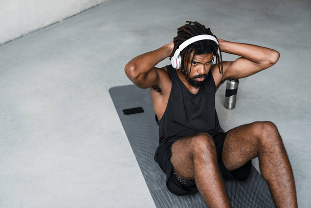 Image of strong african american sportsman in headphones doing exercise while working out on mat indoors - Фото, изображение