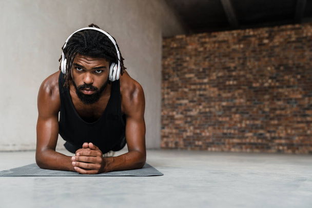 Image of strong african american sportsman in headphones doing exercise while working out on mat indoors - Fotoğraf, Görsel