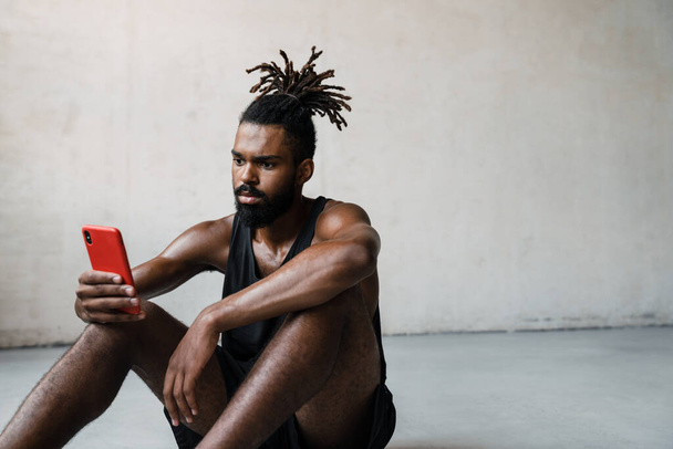 Image of young african american sportsman using mobile phone while sitting on floor indoors - Foto, Imagen