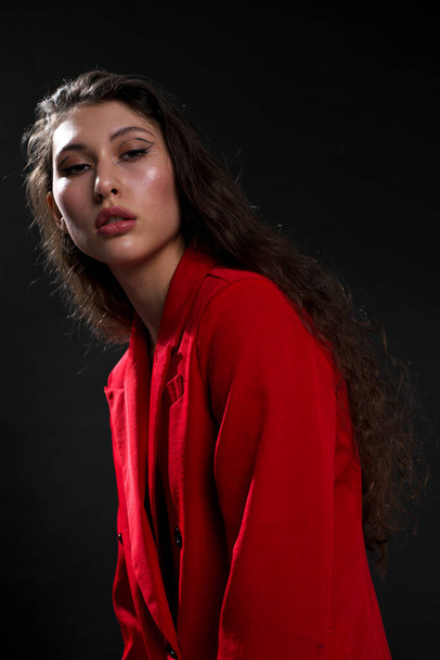 A young beautiful oriental woman with long black hair in a red jacket against a black background. The fashion model poses in the studio. - Foto, immagini