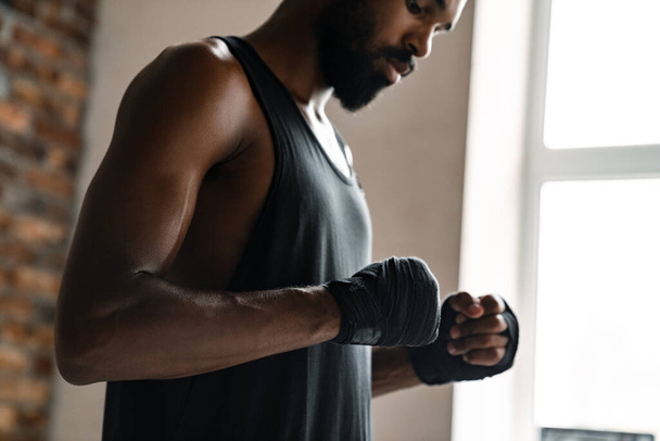 Beeld van atletische Afrikaanse Amerikaanse sporter boksen in de hand verband tijdens het sporten binnen - Foto, afbeelding