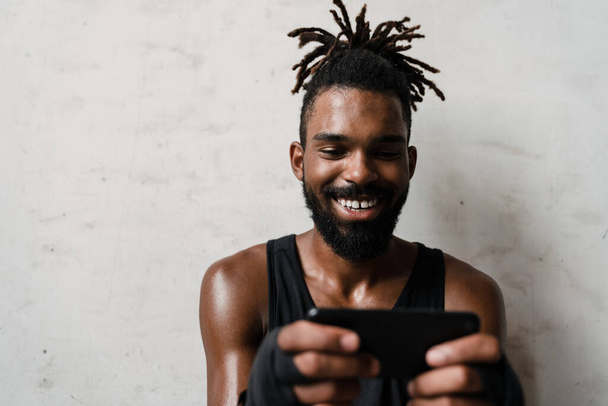 Image of smiling african american sportsman using mobile phone while standing indoors - Foto, Bild