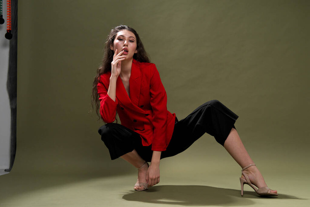 A sensual young woman in a red jacket, culottes and shoes with wet long black hair poses against a green background in the studio. - Photo, image