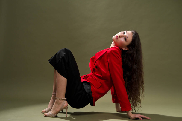 A sensual young woman in a red jacket, culottes and shoes with wet long black hair poses against a green background in the studio. - Foto, Bild