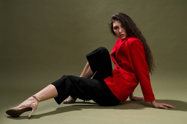 A sensual young woman in a red jacket, culottes and shoes with wet long black hair poses against a green background in the studio. - Zdjęcie, obraz