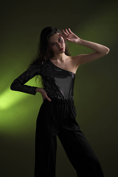 A beautiful young tall girl with long black hair poses in a shiny blouse and black culottes on a green background. Buty and fashion photo shoot. - Valokuva, kuva