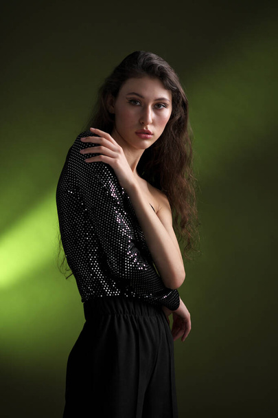 A beautiful young tall girl with long black hair poses in a shiny blouse and black culottes on a green background. Buty and fashion photo shoot. - Foto, imagen