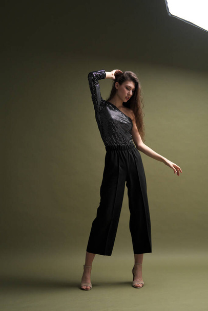 A beautiful young tall girl with long black hair poses in a shiny blouse and black culottes on a green background. Buty and fashion photo shoot. - Φωτογραφία, εικόνα