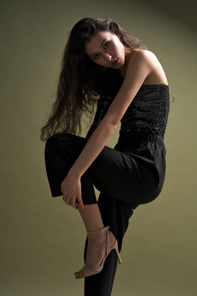 A beautiful young tall girl with long black hair poses in a shiny blouse and black culottes on a green background. Buty and fashion photo shoot. - Φωτογραφία, εικόνα