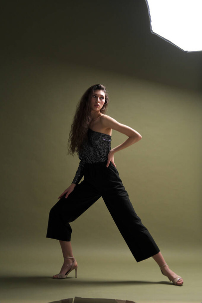 A beautiful young tall girl with long black hair poses in a shiny blouse and black culottes on a green background. Buty and fashion photo shoot. - Fotografie, Obrázek