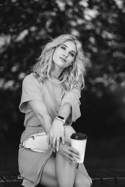 Young beautiful blonde girl in a blue short dress and with a white belt bag walks in a green park. He sits on a bench and drinks takeaway coffee. - Foto, Bild