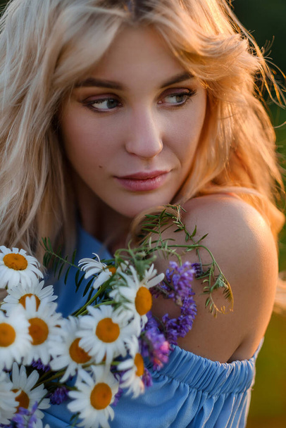 Portrait of a young beautiful blonde girl in a blue dress. Loose hair, sensual lips and big blue eyes. A woman holds a bouquet of daisies wildflowers . - Foto, immagini