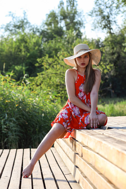 Een mooi meisje zit op een houten brug. Ze is in de buurt van de rivier, gekleed in oranje bloem patroon jurk. Het is zomer zonsondergang. Ze heeft haar hoed op. - Foto, afbeelding