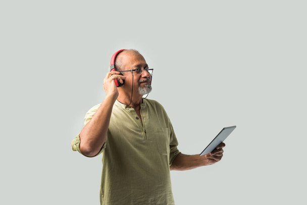 felice anziano indiano asiatico barbuto uomo sorridente utilizzando cuffie con smartphone o tablet su sfondo bianco, presentando schermo o ballare - Foto, immagini