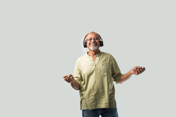 felice anziano indiano asiatico barbuto uomo sorridente utilizzando cuffie con smartphone o tablet su sfondo bianco, presentando schermo o ballare - Foto, immagini
