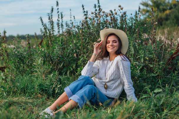 Young beautiful brunette woman dressed in a white sweater, jeans and cowboy straw hat sitting on green grass, smiling and laughing during sunset - Фото, изображение