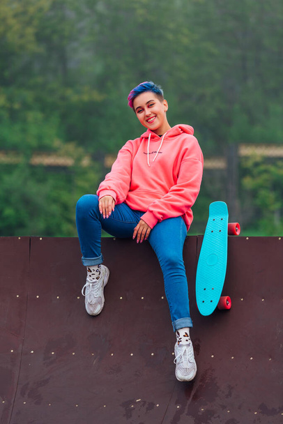 Portrait of a trendy pretty young girl with short colored hair and nose piercing dressed in pink hoodie and jeans sitting next to the skateboard court with her blue plastic skateboard. - Φωτογραφία, εικόνα