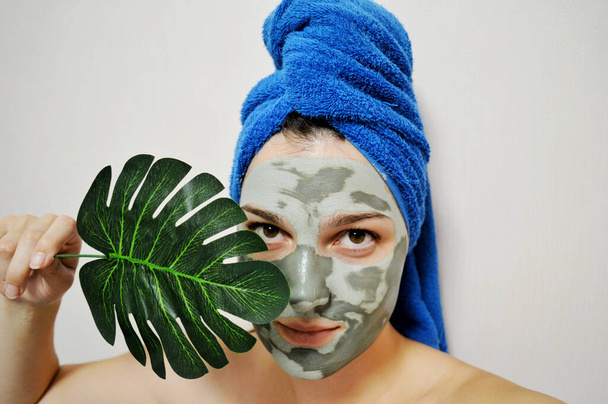 Donna con un asciugamano blu sulla testa e una maschera di argilla sul viso tiene una foglia di palma - Foto, immagini