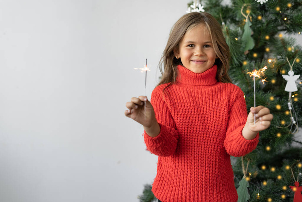 a girl of European appearance is dressed in a red sweater. stands near a Christmas tree with sparklers. Copy space - Фото, изображение