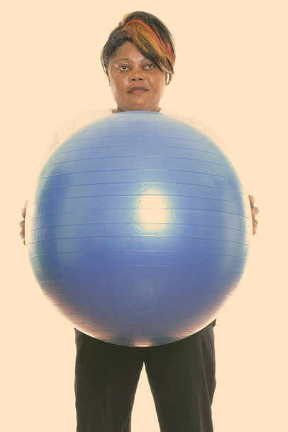Studio shot of fat black African woman thinking and standing while holding exercise ball ready for gym - Foto, imagen